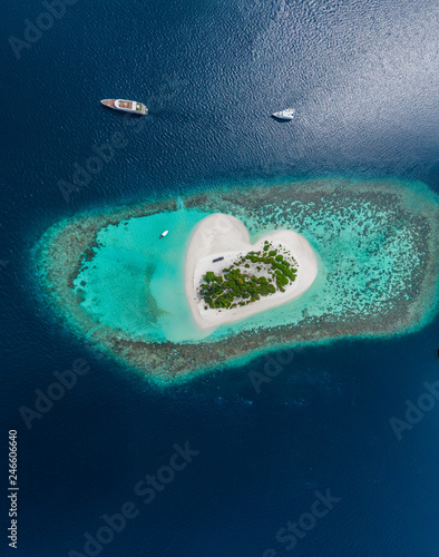 Drone Aerial view of beautiful tropical heart shaped island among blue sea water at Maldives paradise with yachts on panoramic background