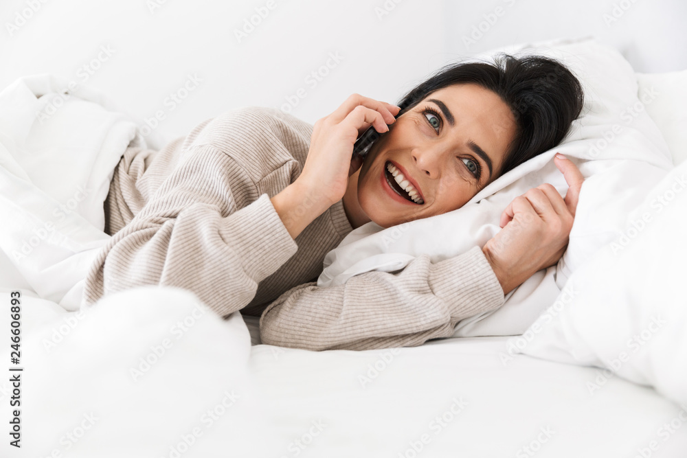 Image of smiling woman 30s using smartphone, while lying in bed with white linen at home Image of smiling woman 30s using smartphone, while lying in bed with white linen at home