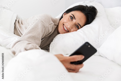Photo of pleased woman 30s using smartphone, while lying in bed with white linen at home