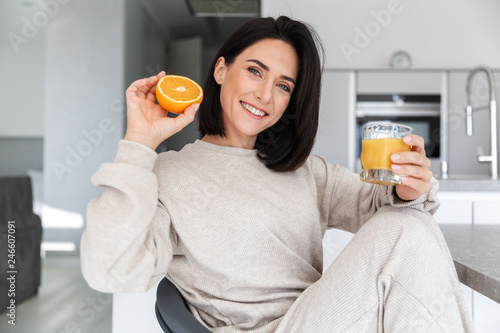 Image of brunette woman 30s drinking orange juice, while resting in bright modern room