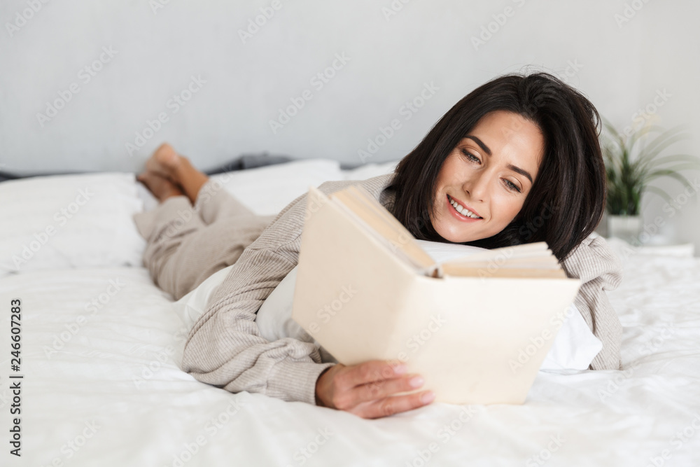 Photo of beautiful woman 30s reading book, while lying in bed with white linen at home Photo of beautiful woman 30s reading book, while lying in bed with white linen at home
