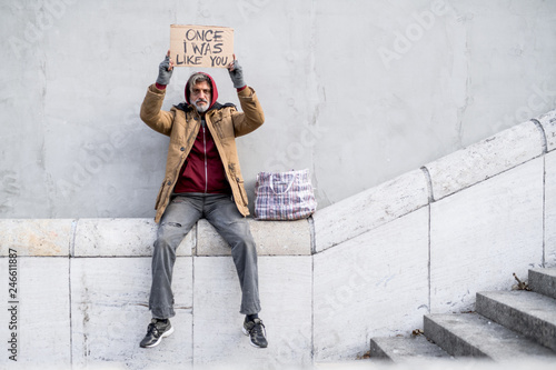 Homeless beggar man sitting in city holding cardboard sign. Copy space.