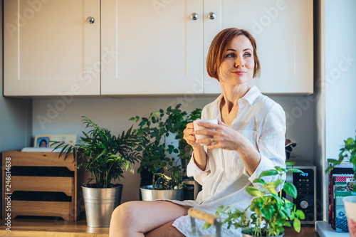 A young woman sitting on worktop indoors in kitchen, holding a cup of coffee.