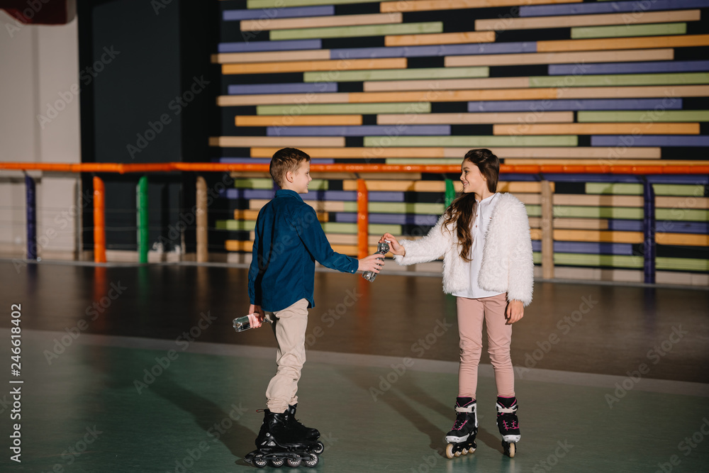 Pretty boy giving to beautiful friend bottle of water, standing on spacious roller rink