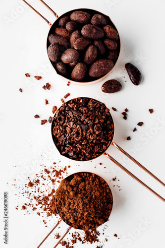 Rose gold measuring cups of cocoa beans, cacao nips and cocoa powder on a white background, flat lay healthy food concept