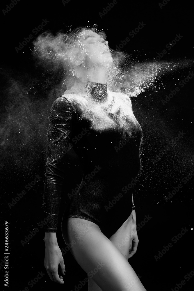 Beautiful slim girl wearing a gymnastic bodysuit covered with clouds of the flying white powder poses on a dark background. Artistic conceptual and advertising black and white photo.