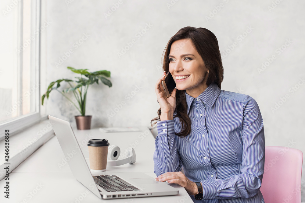 Business woman using laptop and talking on smart phone. Young beautiful joyful girl working on computer Business woman using laptop and talking on smart phone. Young beautiful joyful girl working on computer