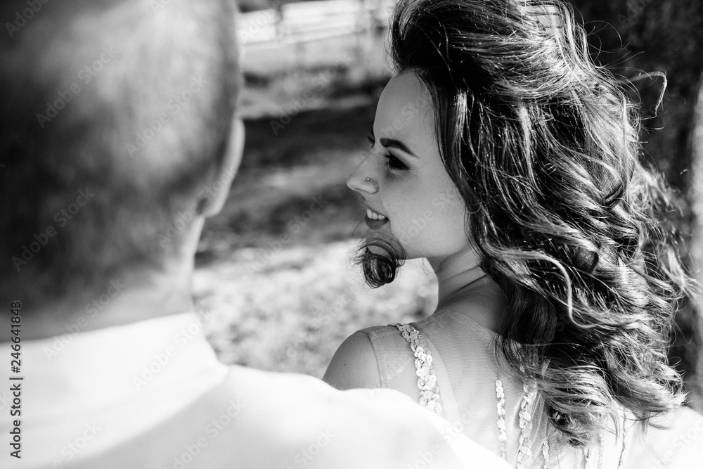 Obraz premium Wedding at sunset. Couple is sitting on a bench under a tree. Beige dress with sparkles. Light suit with a bow tie. The bride and groom embrace and kiss.