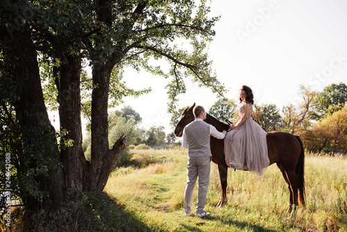 Wedding at sunset. Couple is sitting on a bench under a tree. Beige dress with sparkles. Light suit with a bow tie. The bride and groom embrace and kiss. © malysheva