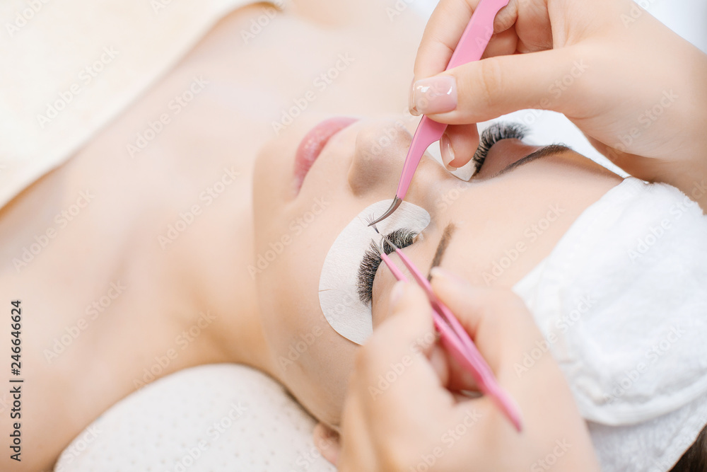 Eyelash Extension Procedure. Close up view of beautiful Woman with Long Eyelashes. Stylist holding pink tweezers, tongs and making lengthening lashes for girl in a beauty salon. Beauty Concept.