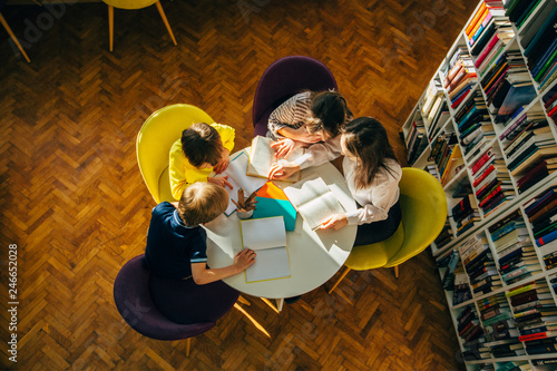 Teacher and children reading fairy tales while sitting in a circle round table at library.Top view of librarian sitting with three multiethnic children. Teacher reading book to girl and boys at school photo