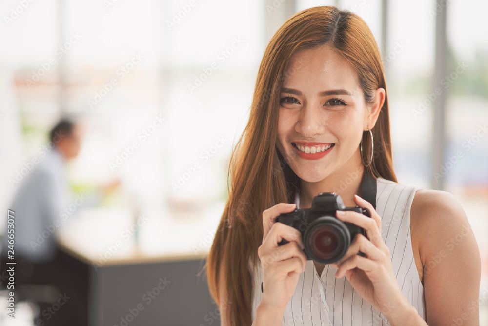 Successful business young Asian beautiful woman looking at camera and hold a camera while colleague discuss or meeting behind her in the office