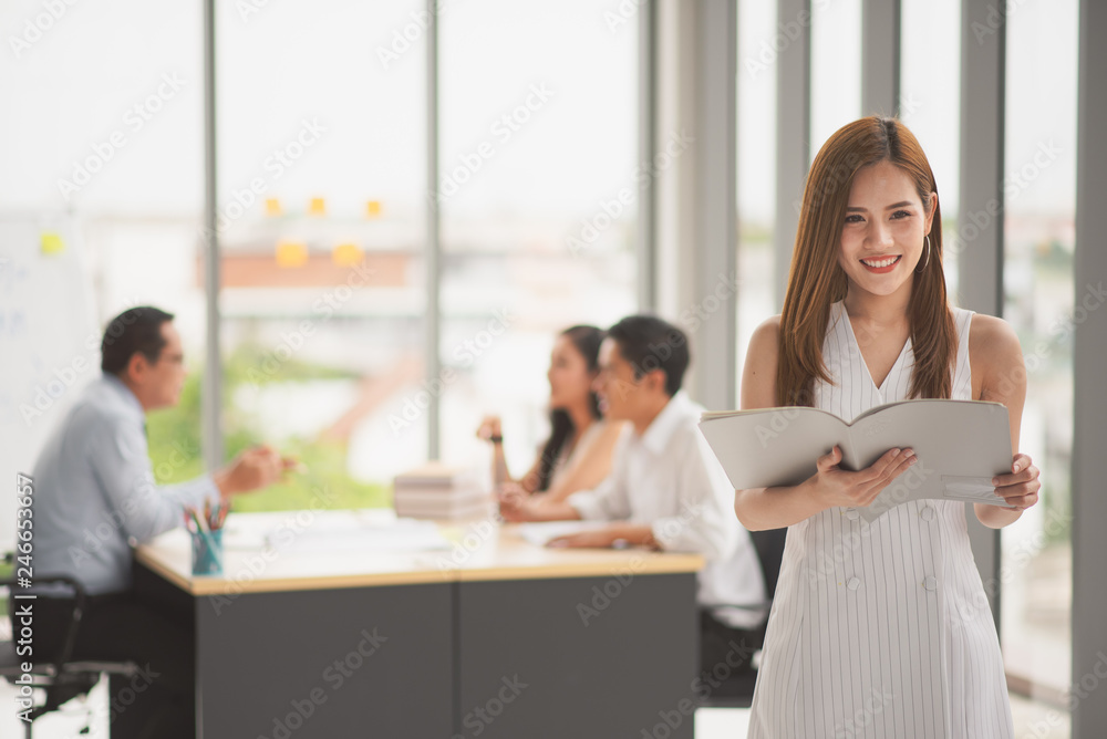 YSuccessful business young Asian beautiful woman looking at camera and hold a book while colleague discuss or meeting behind her in the office