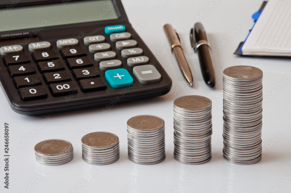 Coin columns, calculator, notepad and pen on a white table Coin columns, calculator, notepad and pen on a white table