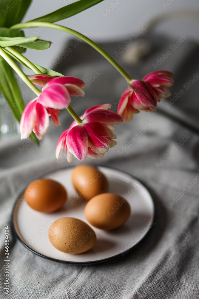 Boiled brown eggs on a plate next to the tulips in a transparent glass cup on a gray background in a muffled light and in soft focus Boiled brown eggs on a plate next to the tulips in a transparent glass cup on a gray background in a muffled light and in soft focus