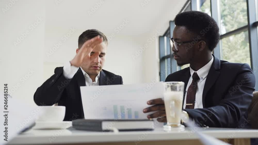 Two businessmen, afro American and caucasian nationality, in business jackets sitting at the office table and discuss the new comercial project, review the papers with graphs. International meeting of Two businessmen, afro American and caucasian nationality, in business jackets sitting at the office table and discuss the new comercial project, review the papers with graphs. International meeting of