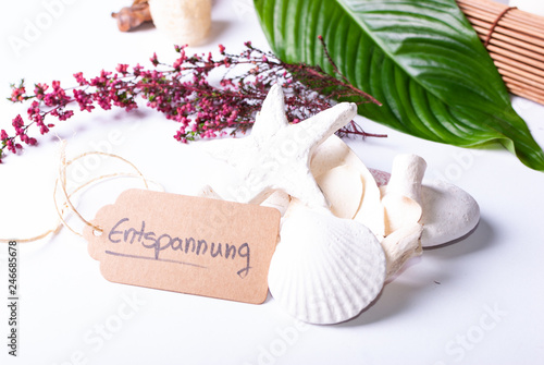 A set up of wellness items, a bamboo mat, a shell, candles, a branch, wood, a starfish and a sign saying "'Relaxation" in German