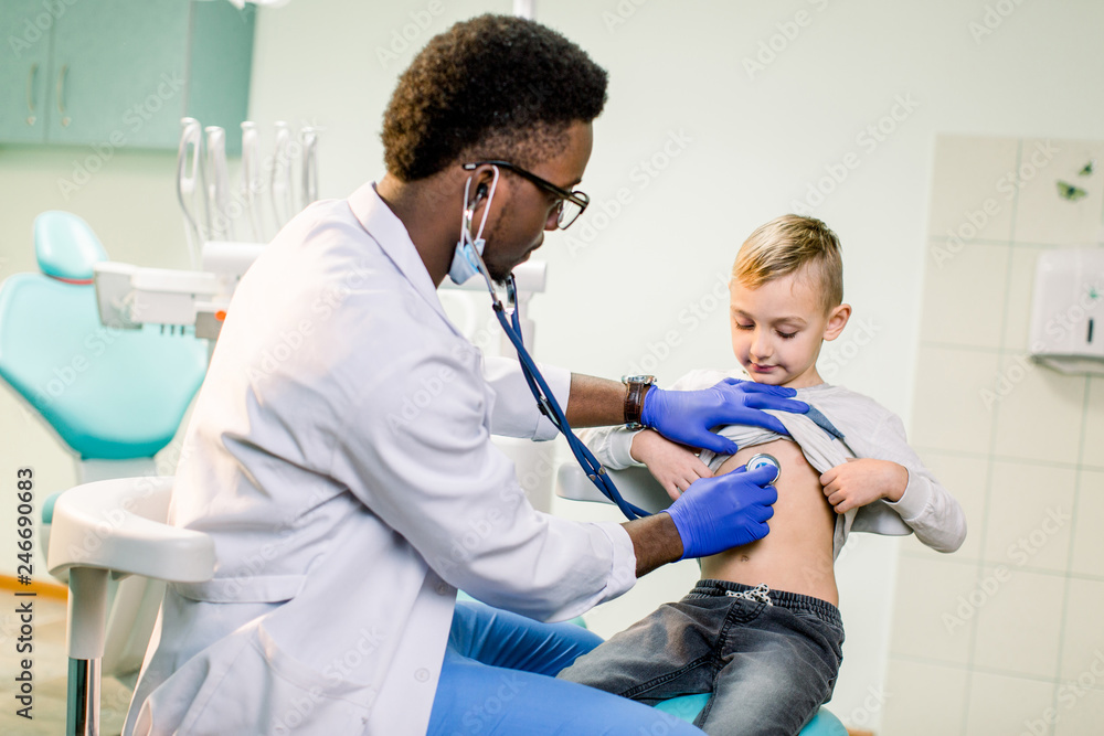 Obraz premium Physician looking at patient and examining posture of little boy. Happy kid smiling, looking away. African American Doctor having stethoscope on neck, wearing in white medical gown