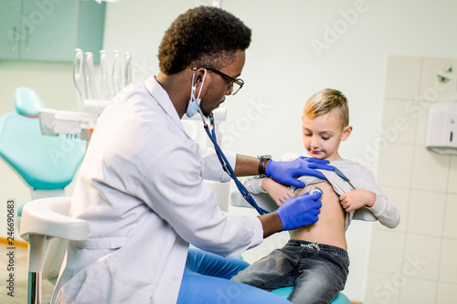 Physician looking at patient and examining posture of little boy. Happy kid smiling  looking away. African American Doctor having stethoscope on neck  wearing in white medical gown