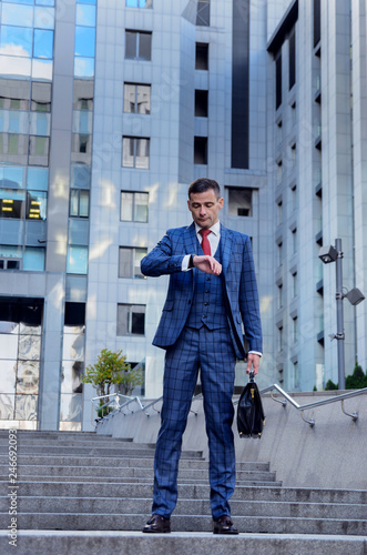 Charismatic man in a plaid suit on the background of the business center (office). Looks at his watch, checks time, is late, hurries to a business meeting