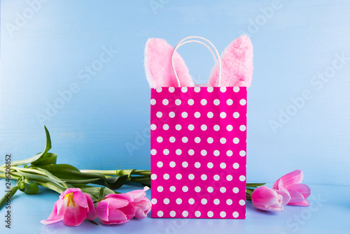 Toy Easter bunny ears sticking out of spotted paper shopping bag with pink tulips on blue background.