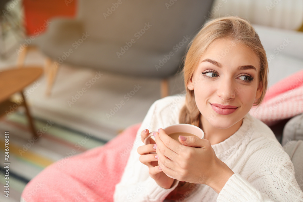 Beautiful young woman wrapped in plaid sitting on sofa with cup at home. Winter atmosphere Beautiful young woman wrapped in plaid sitting on sofa with cup at home. Winter atmosphere