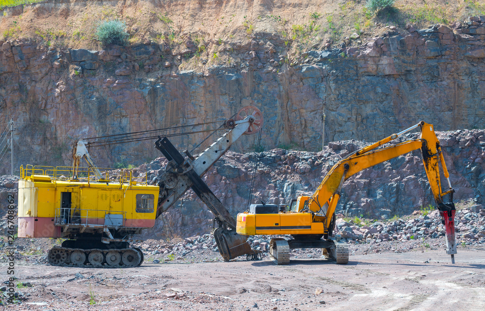 A large tracked excavator and pneumatic hammer in a quarry open pit mining of granite stone. Process production stone and gravel. Quarry mining equipment. A large tracked excavator and pneumatic hammer in a quarry open pit mining of granite stone. Process production stone and gravel. Quarry mining equipment.