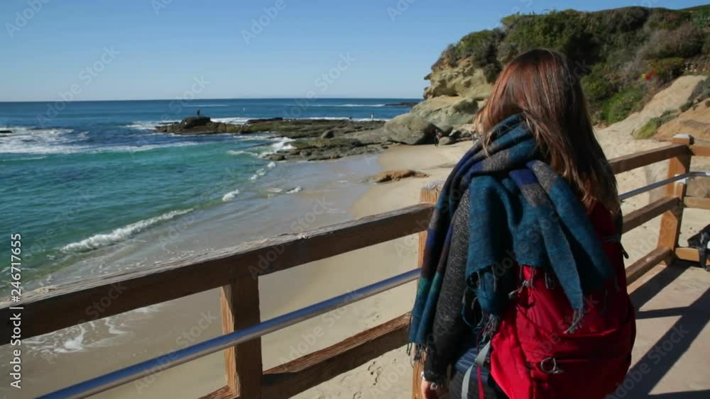 Back view of young woman with backpack and scarf getting closer to the fence on the ocean beach. Back view of young woman with backpack and scarf getting closer to the fence on the ocean beach.