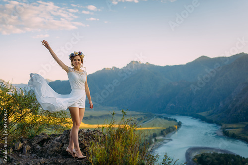 Beautiful bride in white short dress posing over ecological landscape of mountains and river with blue cloudy sky