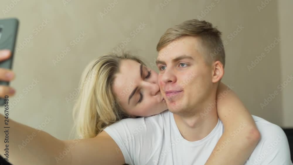 Lovable blonde couple is sitting on a bed in their flat, hugging and ...