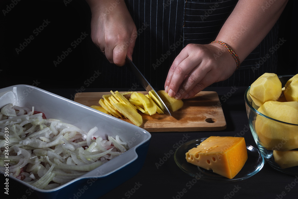 Female chef cutting potato for potato baked pudding with meat on black wooden table, hands, close up. Selective focus.