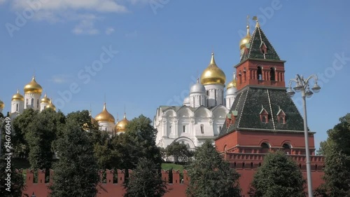 View on red brick Kremlin walls, one of its towers, green trees, and beautiful white cathedrals with golden domes against blue sky in Moscow, Russia
