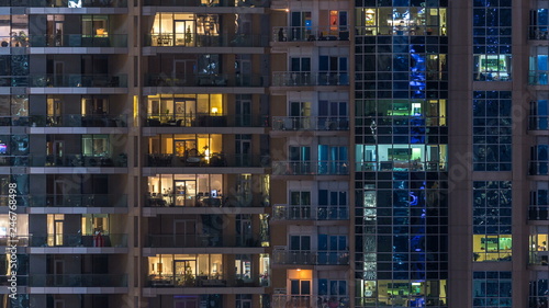 Glowing windows in multistory modern glass residential building light up at night timelapse.
