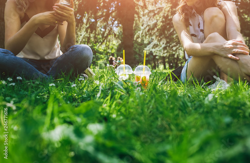 Two hipster girls are sitting on the grass with summer cocktails. Cold non-alcoholic drinks with ice to go. Mojito and strawberry lemonade with lime, soda water and mint. Freshness and holidays.