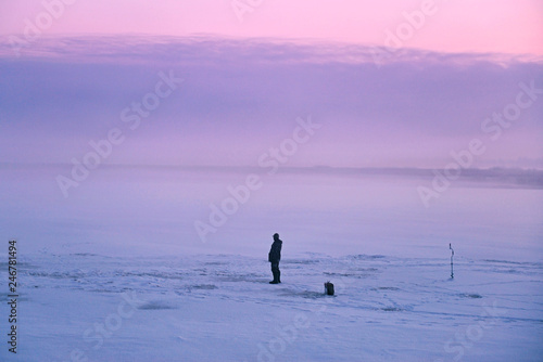 Man on the winter fishing stands on the river ice before sunrise at early morning