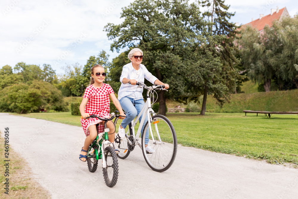 Obraz premium family, leisure and people concept - happy grandmother and granddaughter riding bicycles at summer park