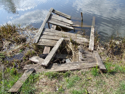 An old wooden destroyed bridge over the river is half under water among green grass 