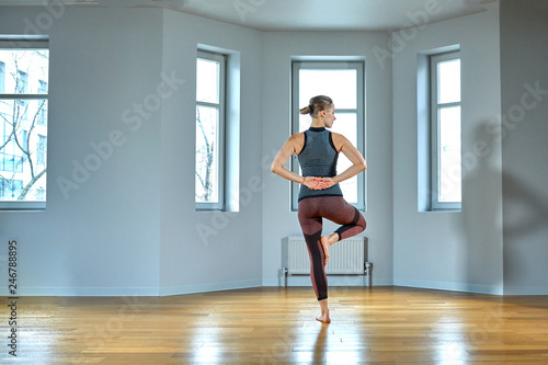Young sporty woman practicing yoga, working out, wearing sportswear, pants and top, indoor close up, yoga studio. Rear view
