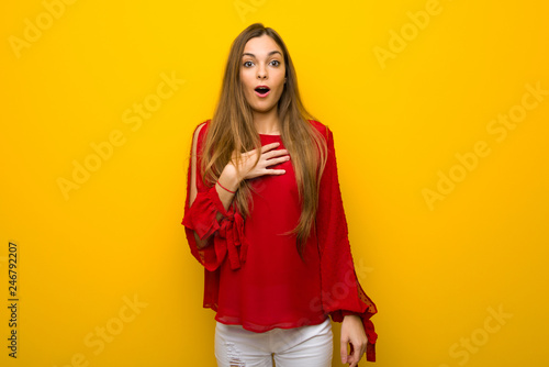 Young girl with red dress over yellow wall surprised and shocked while looking right