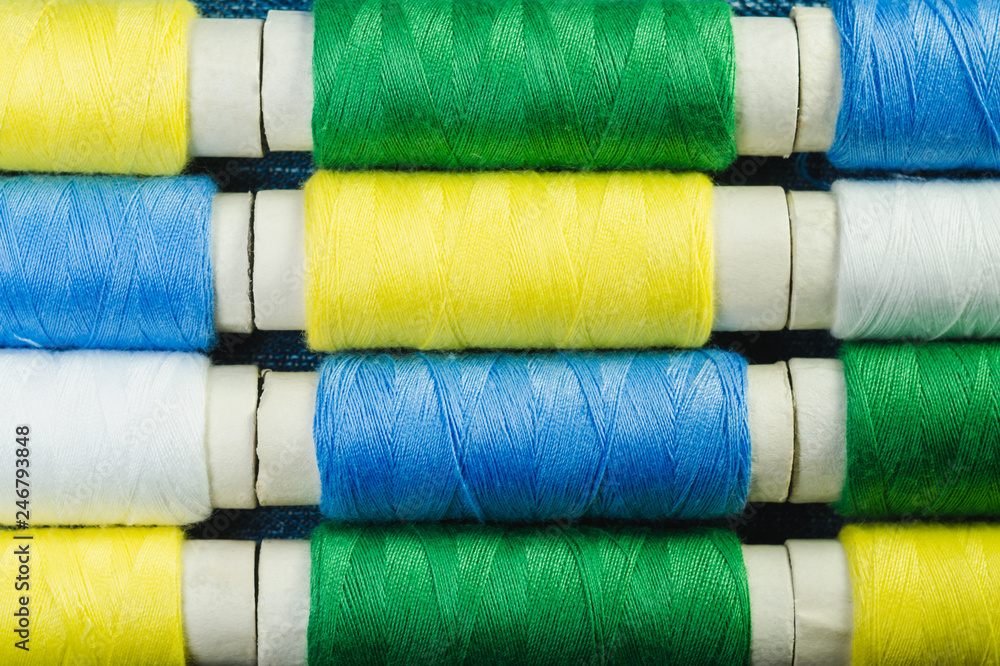 Spools of blue, yellow, green and white sewing thread arranged in rows on denim Spools of blue, yellow, green and white sewing thread arranged in rows on denim