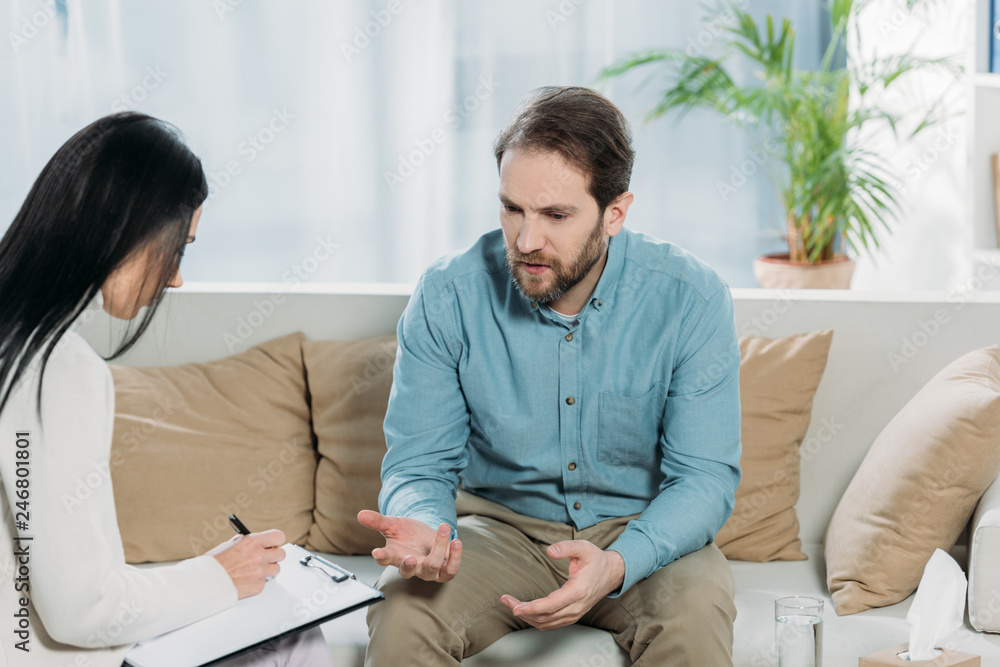 Fototapeta premium psychotherapist writing on clipboard and unhappy bearded man sitting on couch and talking