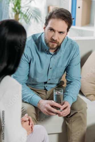 cropped shot of upset bearded man holding glass of water and looking at psychotherapist in office