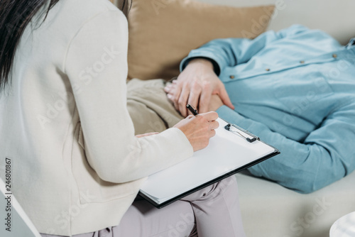 cropped shot of psychotherapist writing on clipboard and male patient lying on couch