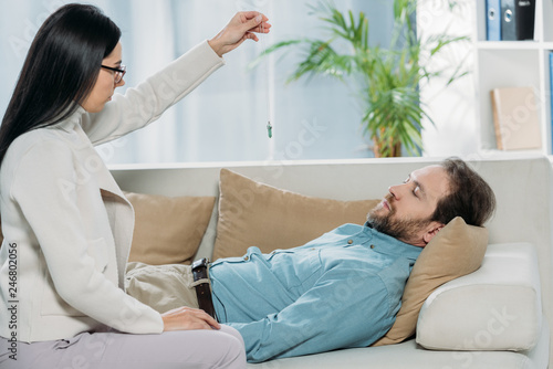 side view of young hypnotist with pendulum and bearded man with closed eyes lying in couch during hypnotherapy photo