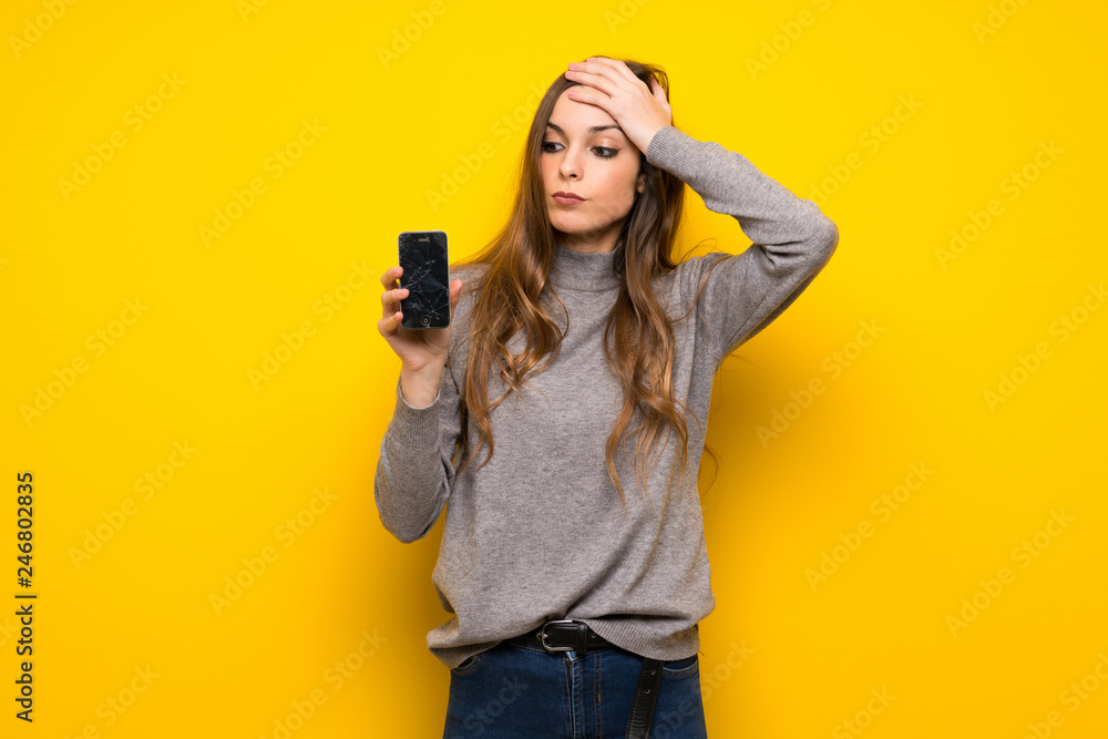 Young woman over yellow wall with troubled holding broken smartphone Young woman over yellow wall with troubled holding broken smartphone