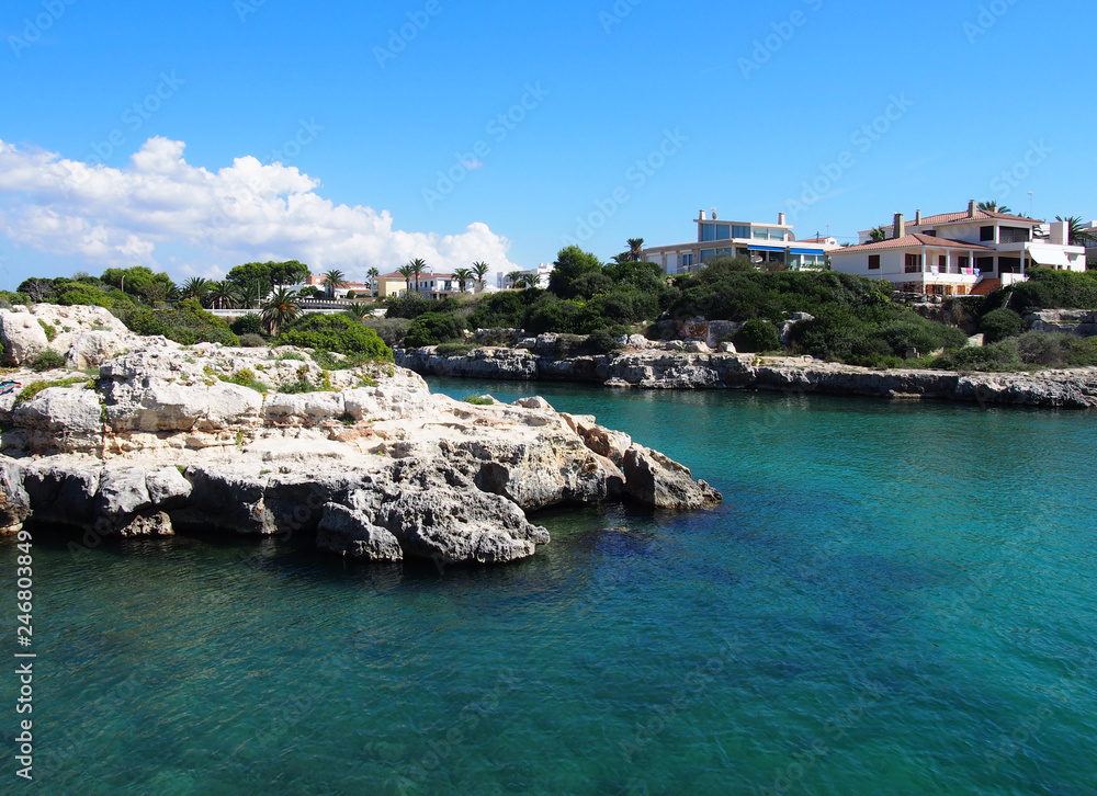 scenic view of the cala santandria in menorca with bright sunlit sea surrounded by cliffs and white houses overlooking the water