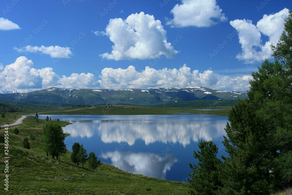 Beautiful sunny day overlooking the lake in the Altai Republic in Russia. White clouds in the blue sky reflected in the water and green trees
