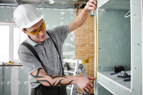 Room repair. Builder measures the cabinet with shelves using a tape measure on the background of the construction site