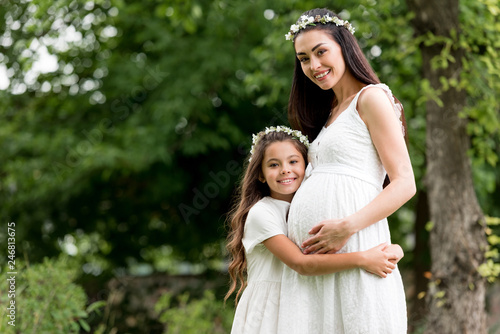 happy child hugging pregnant mother and smiling at camera in park