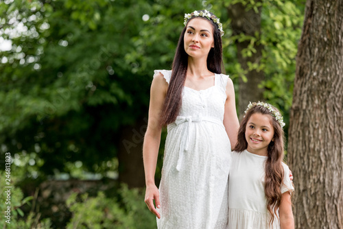 happy pregnant mother and cute daughter in white dresses and wreaths standing together and looking away in park